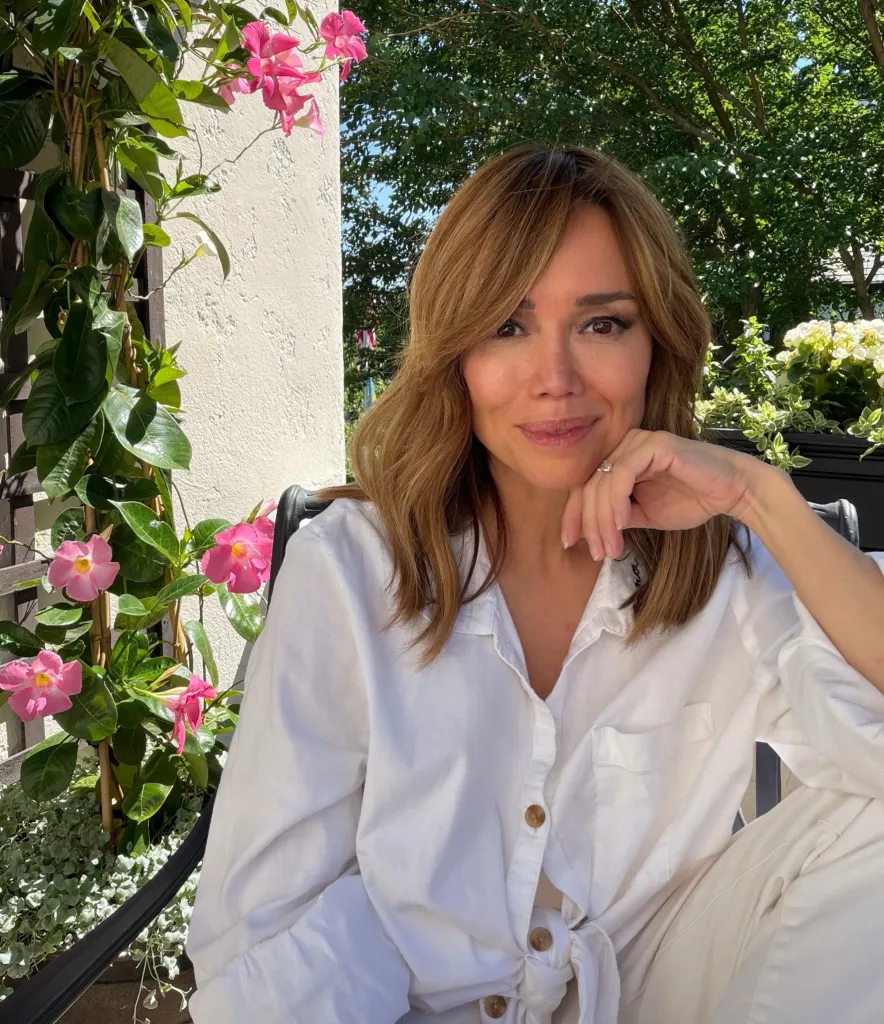 Sara Gore, in a white shirt and pants, sitting outside with pink flowers in the background.