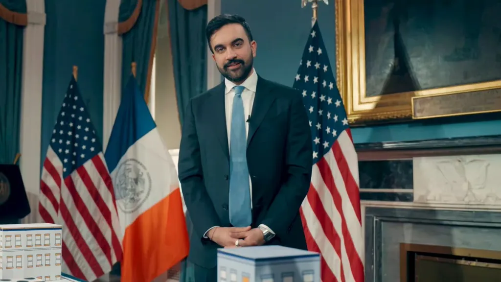 New York City Mayor Zohran Mamdani wears a dark suit, white shirt and blue tie as he stands between two flags.