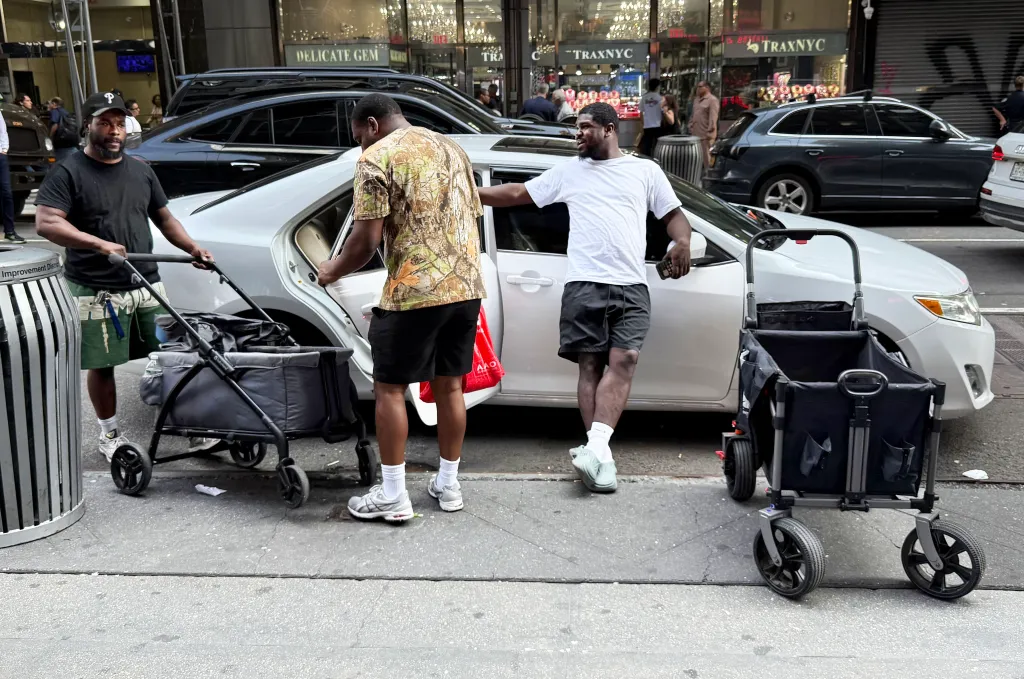 Three men, two pushing empty baby strollers and one leaning on a car, on a street lined with shops and cars.