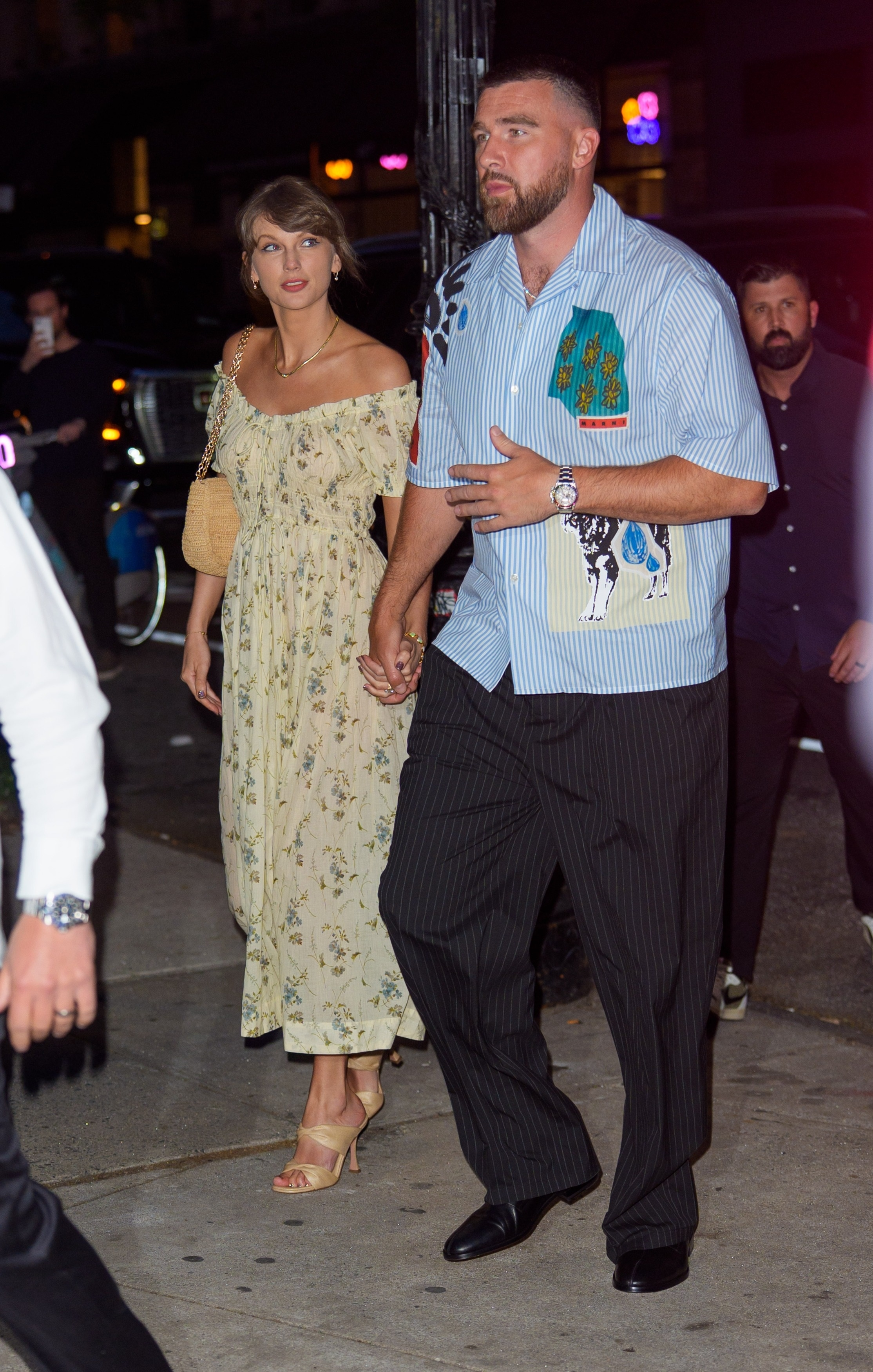 Taylor Swift in a floral dress and Travis Kelce in a striped shirt holding hands during a date night.