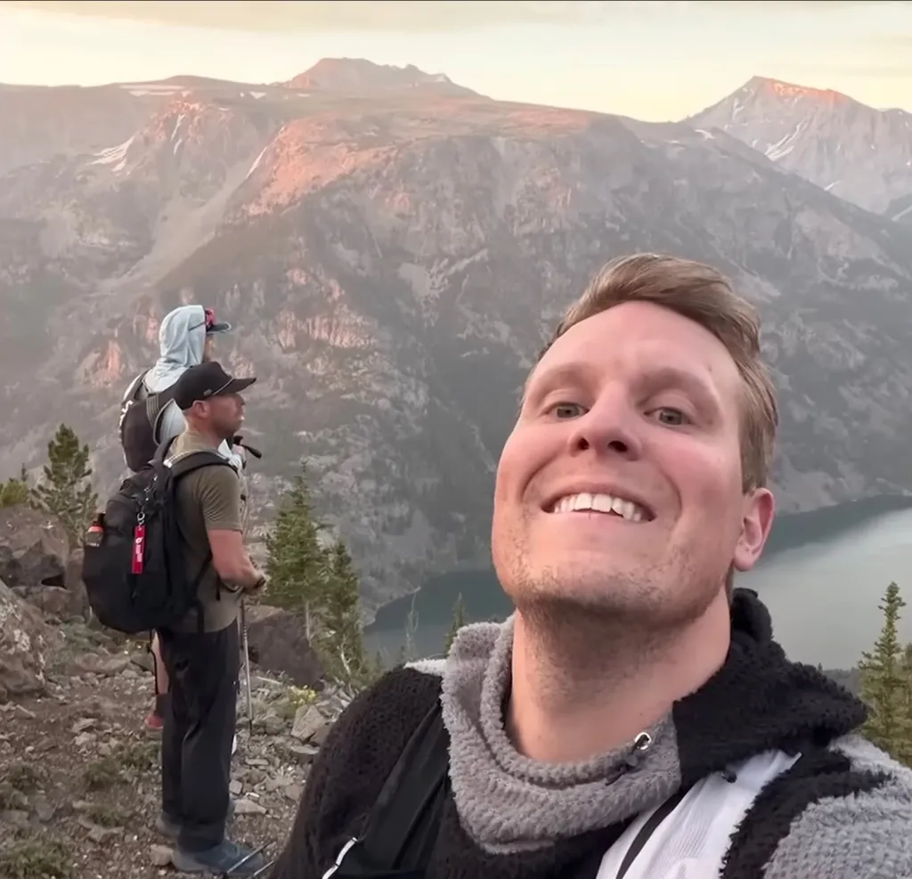A man takes a selfie on a mountain with two other hikers and mountains behind him.