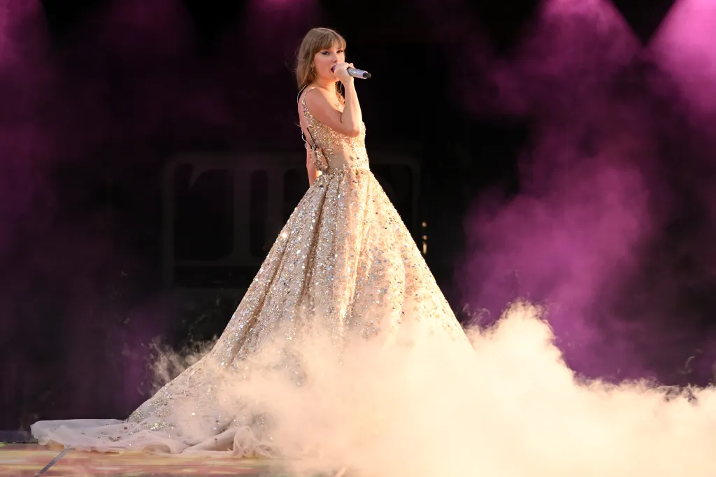 Taylor Swift singing into a microphone on stage, wearing a sparkling golden gown with her dress blending into the stage fog.