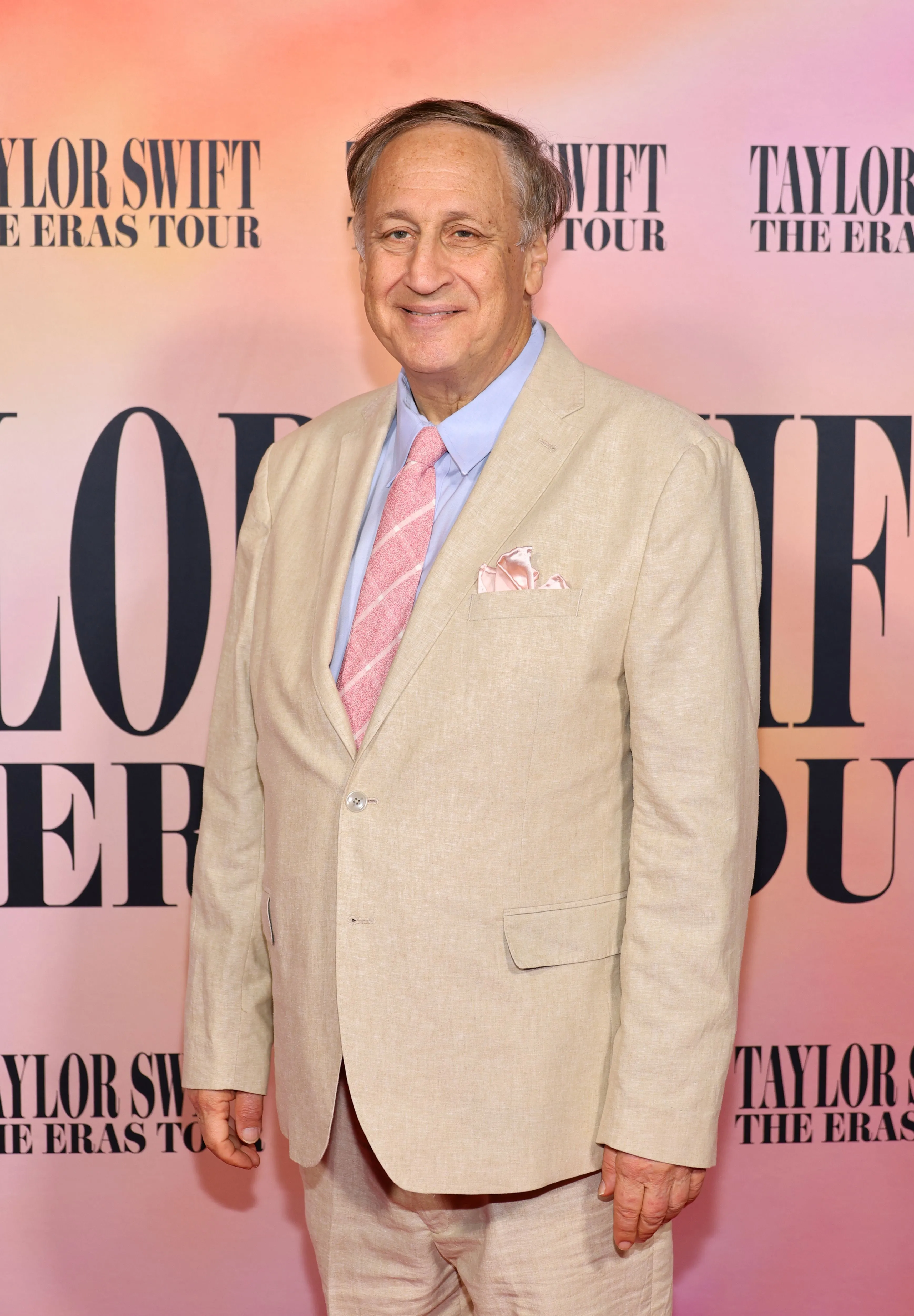 Adam Aron in a tan suit and pink striped tie on a pink background with 