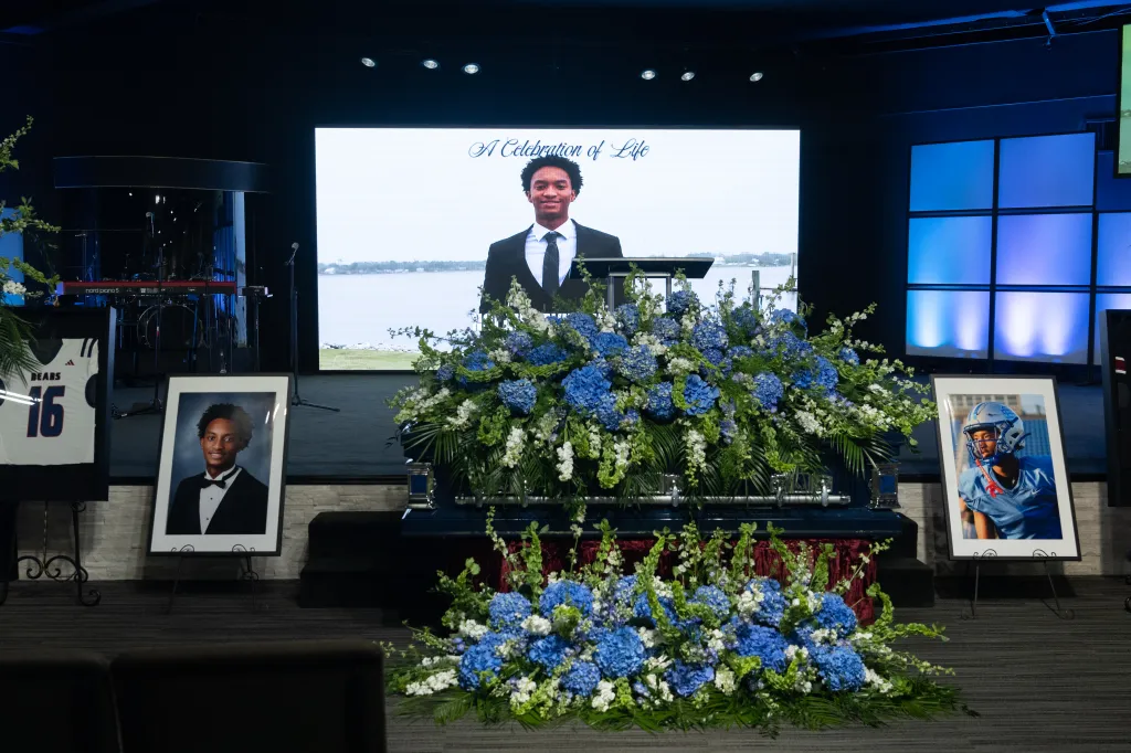 Funeral for Nolan Wells with images of him on screen and in frames, a football jersey, and a casket covered in blue and white flowers.