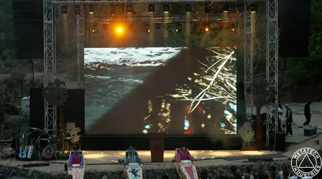 A large outdoor stage with a big screen showing a video, and three people in coffin-like boxes in front.