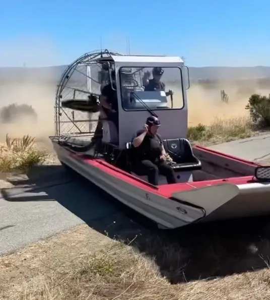 An airboat with two people on board driving off-road, kicking up a large cloud of dust.