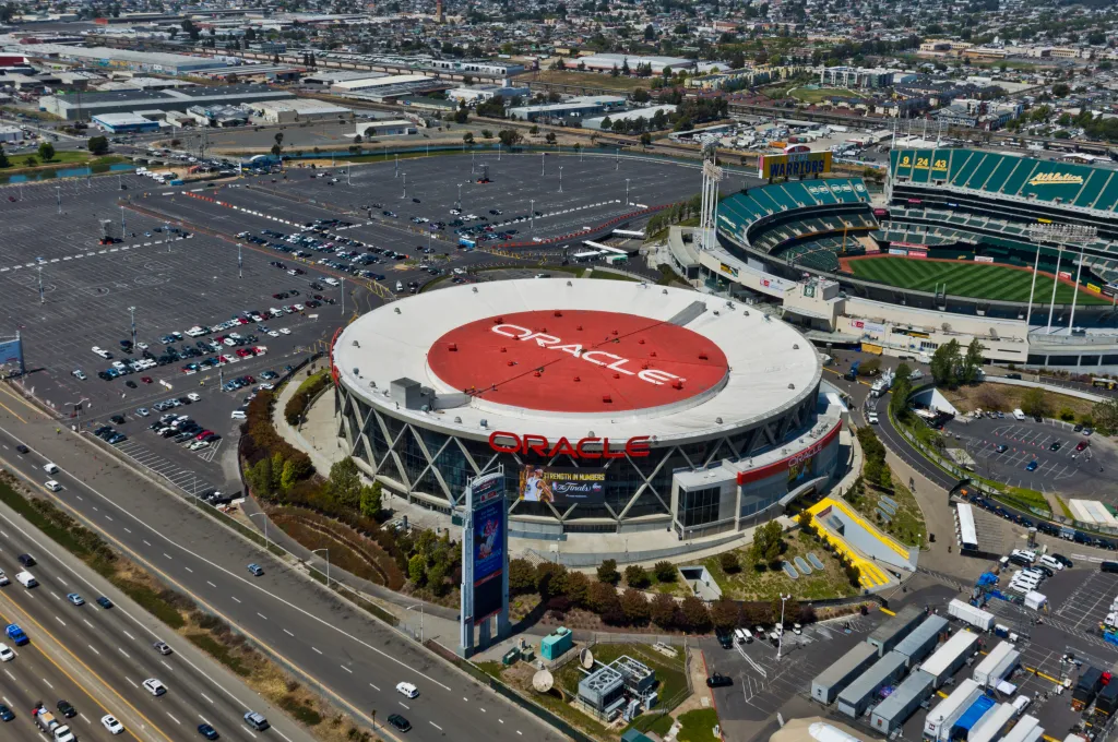 Aerial view of Oracle Arena and a large parking lot, with a highway in the foreground and a stadium in the background.