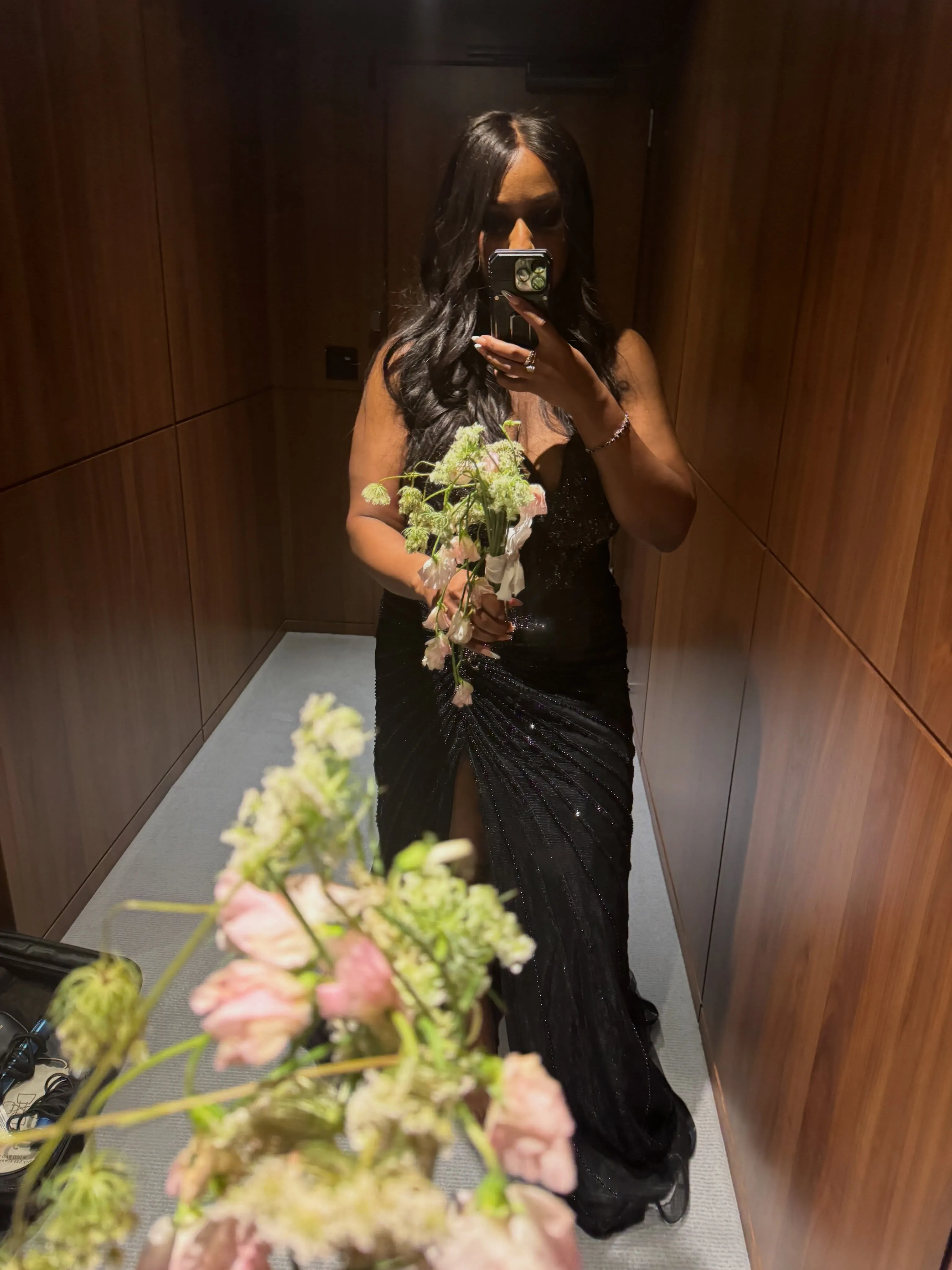 A person in a black gown holds a bouquet of flowers, taking a mirror selfie.