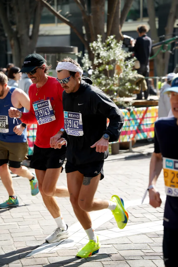 Harry Styles running at the Tokyo Marathon, with other participants, wearing a black jacket, shorts, green sneakers, sunglasses, and a white headband.