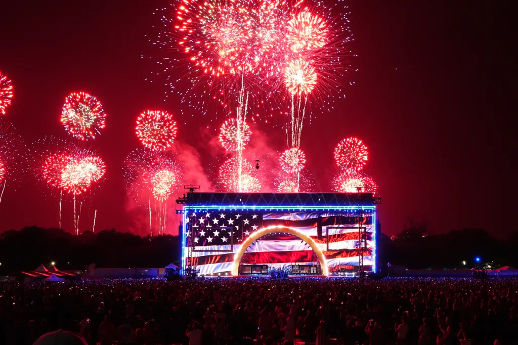 People watch fireworks over a stage decorated with an American flag during an Independence Day event.