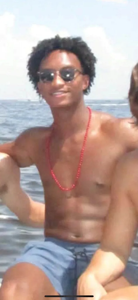 Nolan Xavier Wells, an 18-year-old with dark curly hair and sunglasses, smiles, wearing a red beaded necklace and blue shorts, sitting in the water.