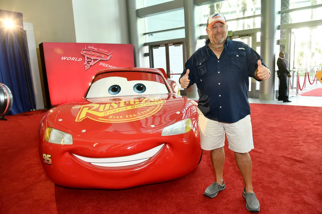 Larry the Cable Guy giving a thumbs up next to a life-size Lightning McQueen car.