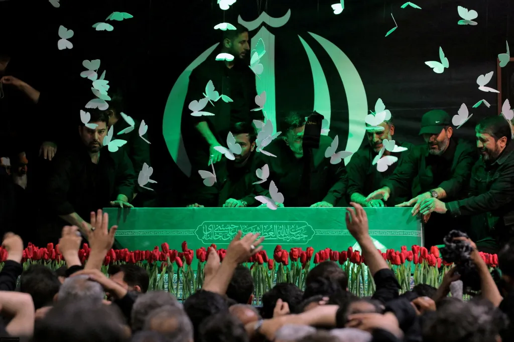 Mourners at a farewell ceremony for Iran's late Supreme Leader, with the green coffin topped with red tulips and surrounded by men in dark clothing.