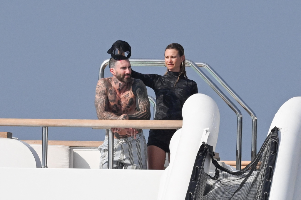 Adam Levine, shirtless and covered in tattoos, with Behati Prinsloo in a black camo-print wetsuit, standing on a yacht in Saint-Tropez.