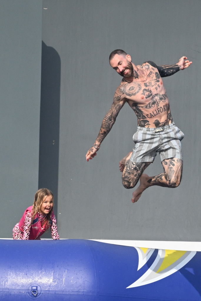 Adam Levine jumping out of the water with his daughter looking up at him and laughing.