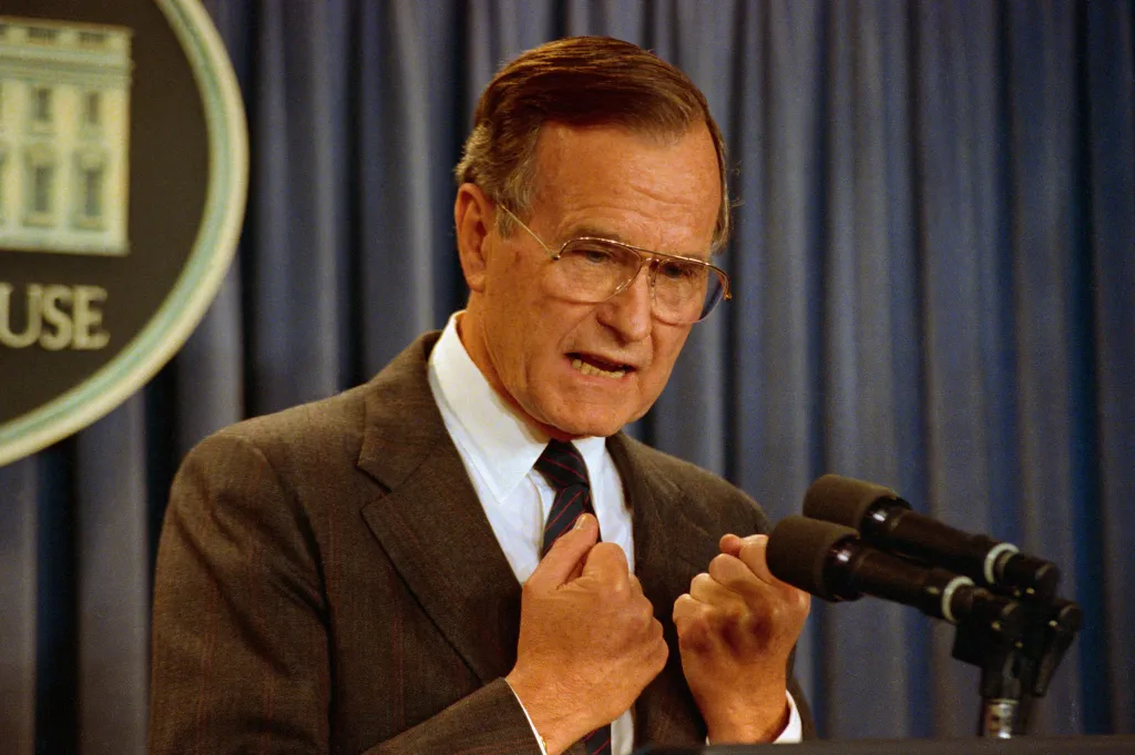 George H.W. Bush speaking at a news conference, with his hands clasped and microphones in front of him.