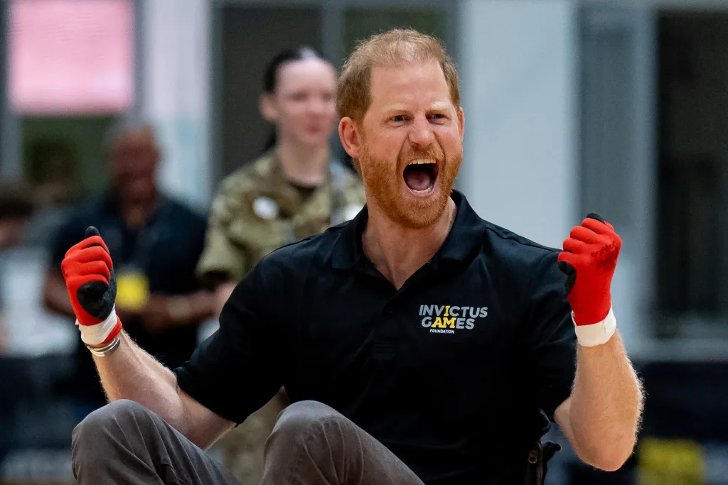 Prince Harry, Duke of Sussex, in a black polo shirt and red gloves, shouts excitedly during a wheelchair rugby game.