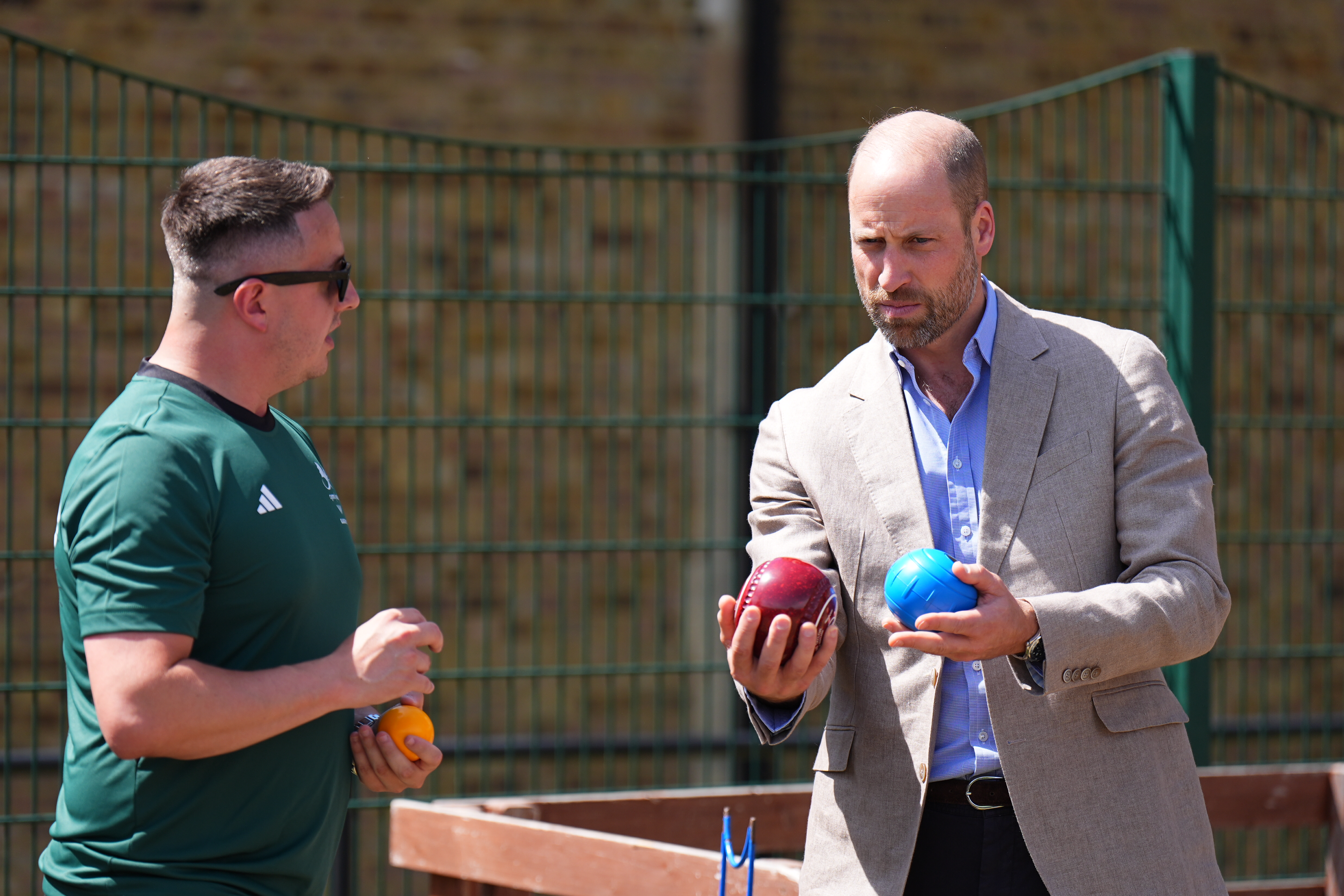 Prince William holding a red and blue bowls ball, looking at a man in a green shirt and sunglasses holding an orange ball.