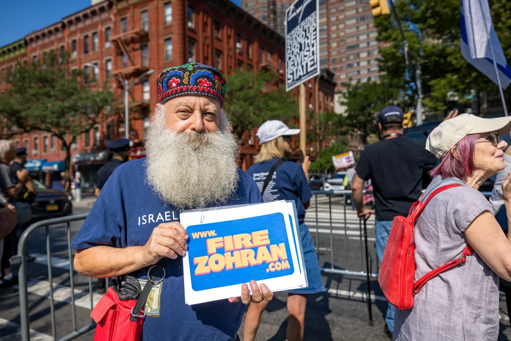 A man wearing a multi-colored embroidered cap and a full white beard holds a 