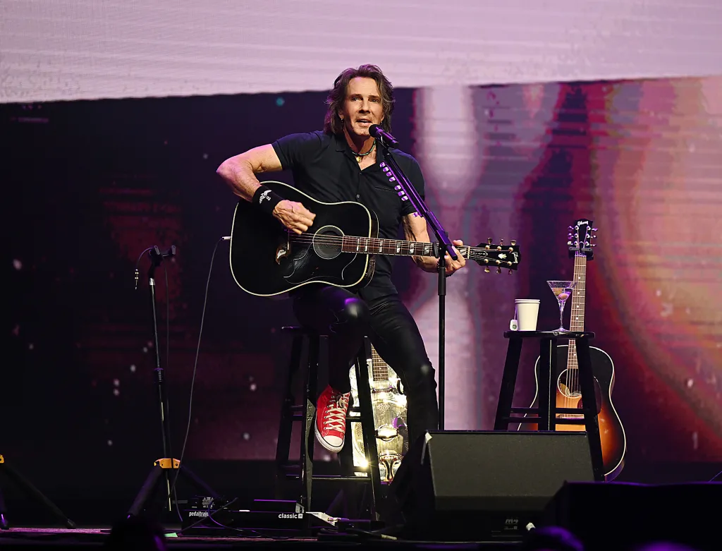 Rick Springfield performing on an acoustic tour, playing a black acoustic guitar while seated on a stool.