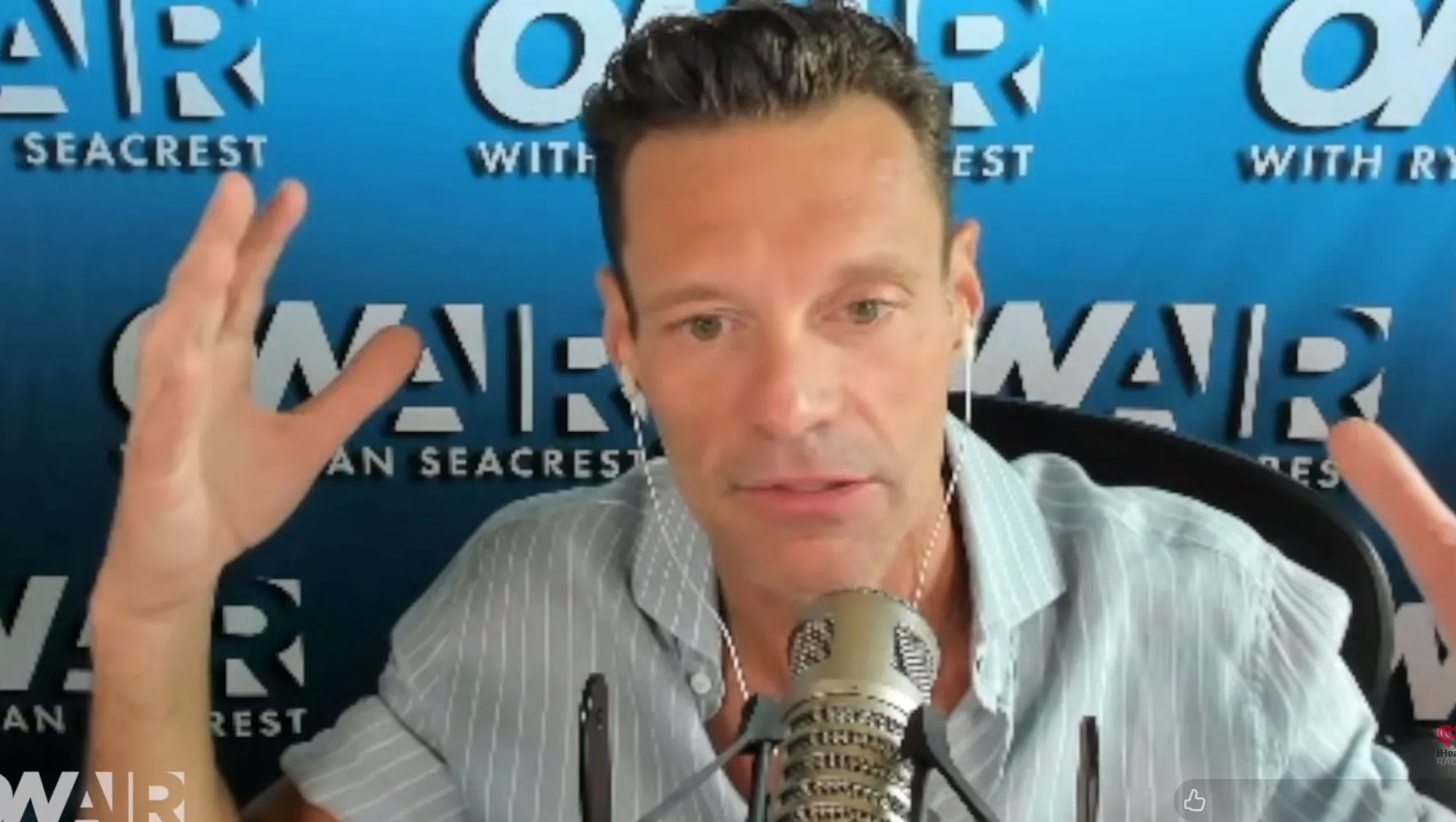 Ryan Seacrest speaking into a microphone with his hands raised, against a blue background with 