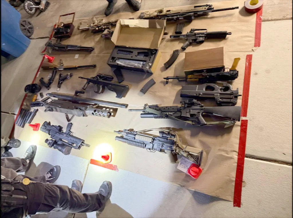 A large assortment of firearms and ammunition laid out on a tarp, with two people's legs visible in the bottom left.