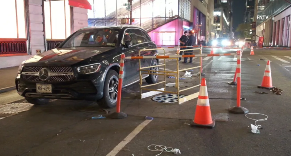 A Mercedes-Benz SUV next to an uncovered manhole surrounded by barriers, cones, and police officers at night.