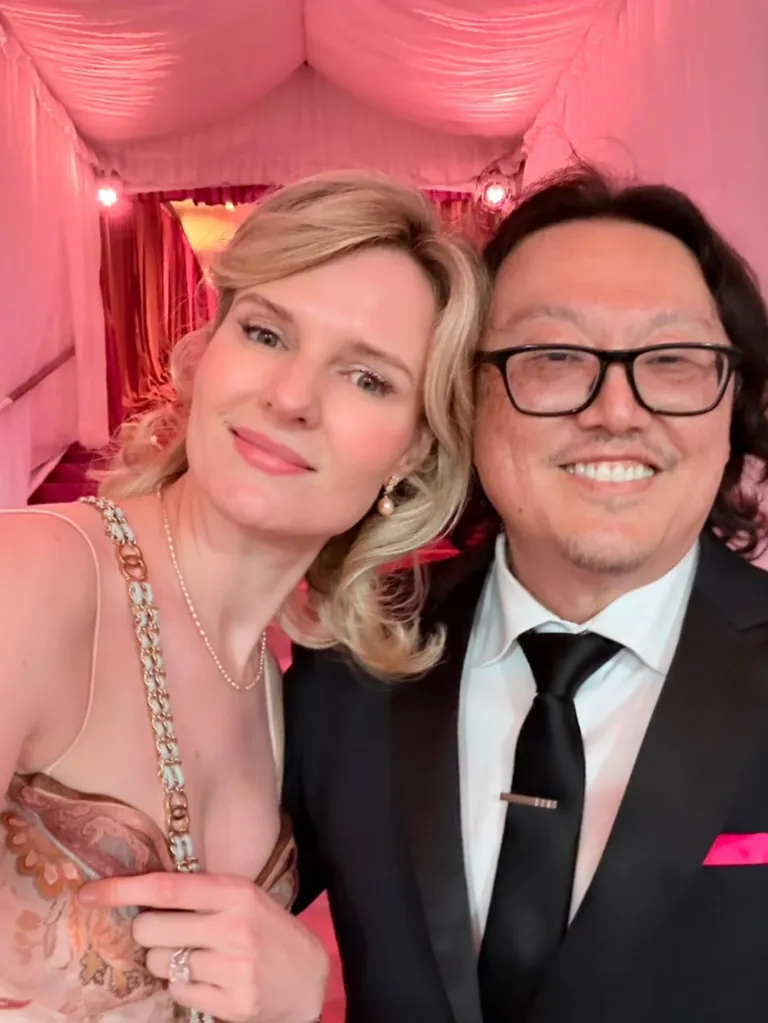 A blonde woman in a patterned dress with a pearl earring and a man in a black suit, white shirt, black tie, and glasses smile for a selfie.