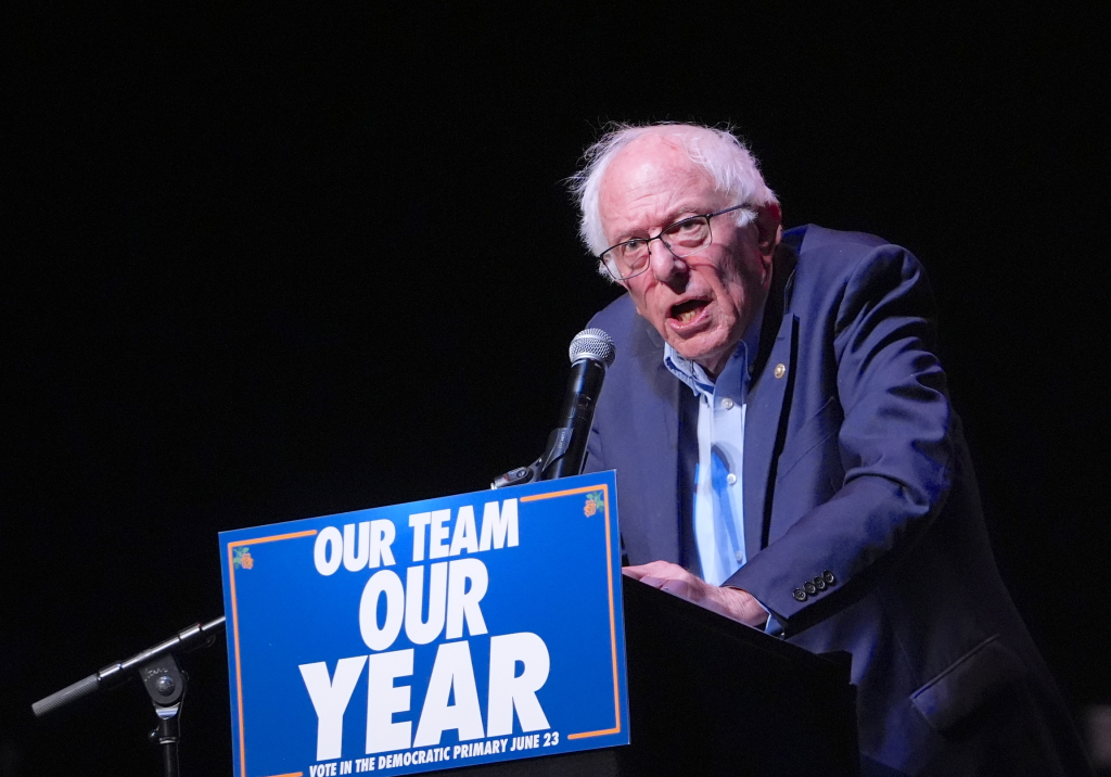 Senator Bernie Sanders speaking at a podium with a microphone, with a blue sign in front that reads 