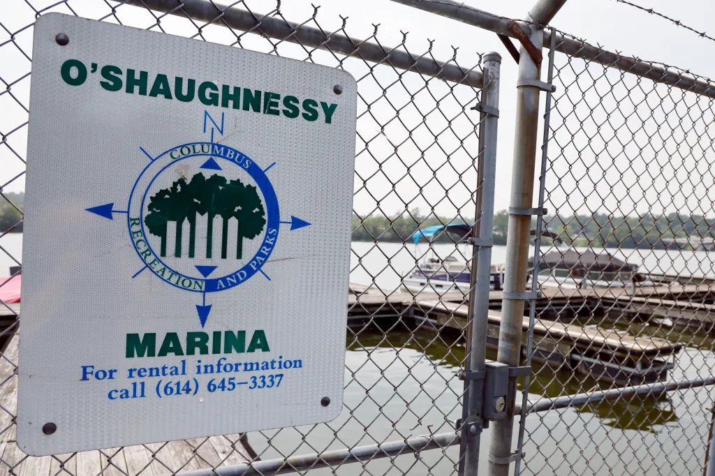 A sign for O'Shaughnessy Marina, Columbus Recreation and Parks, hangs on a chain-link fence overlooking a river with boats docked.