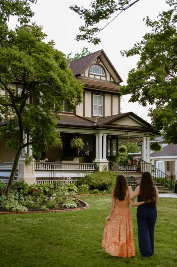 Two women with long brown hair, seen from behind, approach a large, historic home with a wide porch and green landscaping.