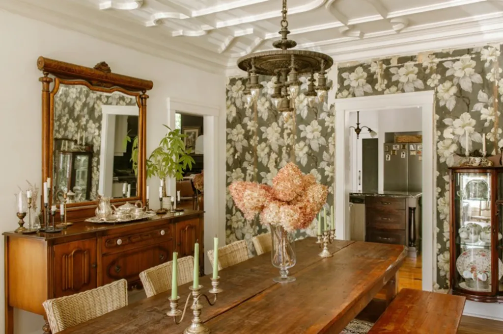 A dining room featuring floral wallpaper, a large wooden table with flowers and candelabras, and an antique sideboard with a mirror.