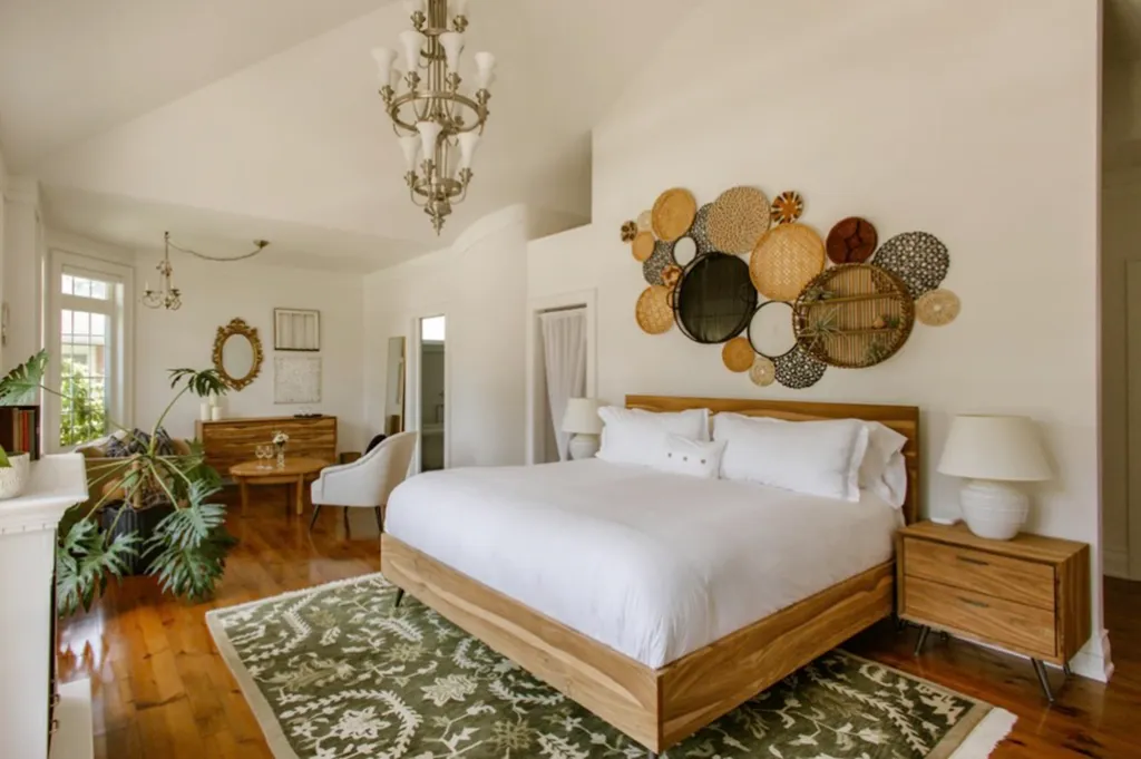 A bright bedroom with a wooden bed, white bedding, a large green rug, a sitting area, and woven wall art above the bed.