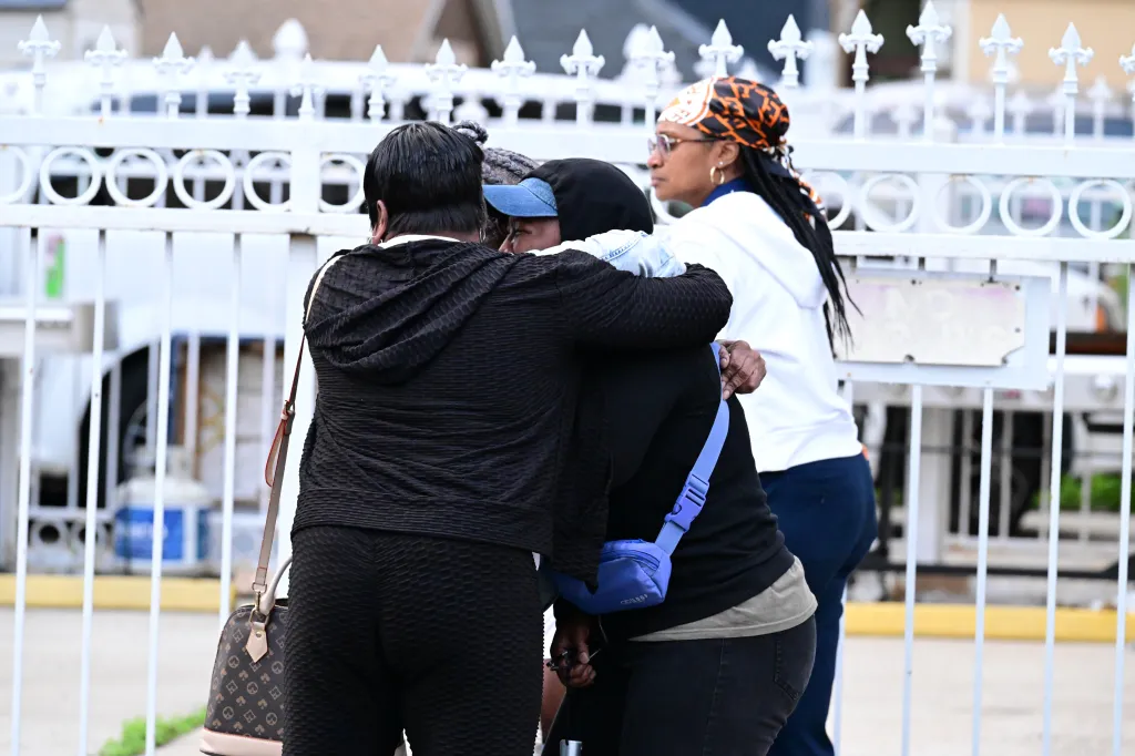 Two women embrace near a white fence, with a third woman standing to the right.