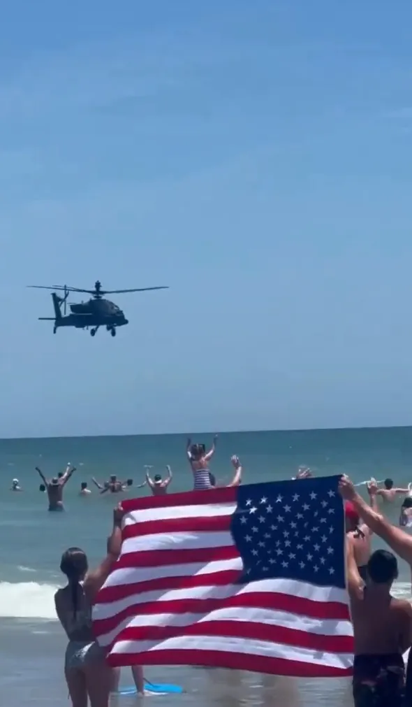 People on a beach in the water holding an American flag as a helicopter flies overhead.