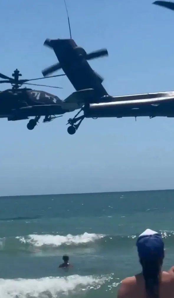 Two helicopters flying low over a beach where people are swimming in the ocean.