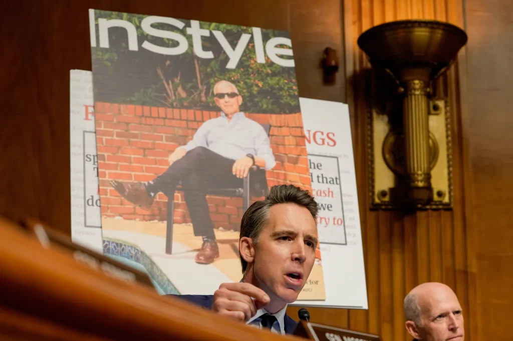 Senator Josh Hawley speaking, with a poster board behind him displaying a magazine cover of Anthony Fauci in a chair with the title 