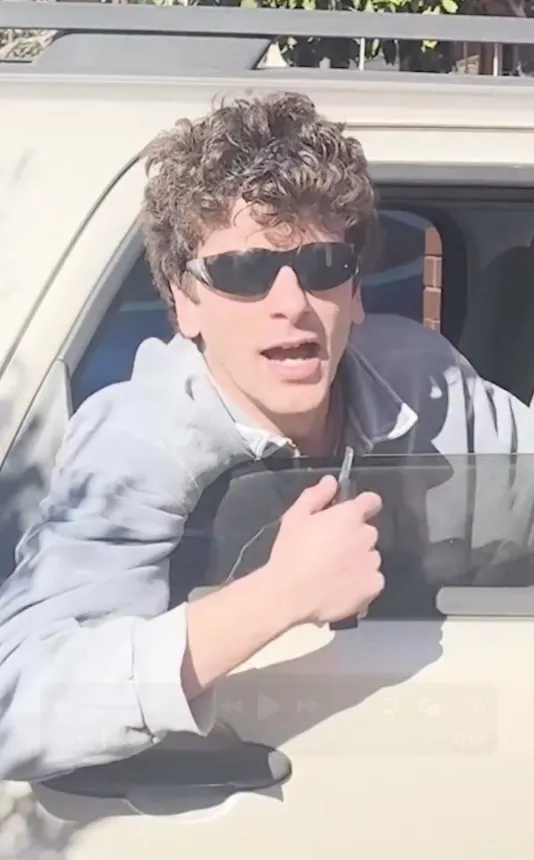 A man with curly hair and sunglasses leans out of a moving car, speaking.