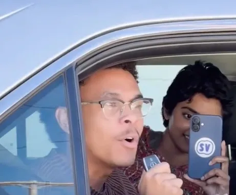 Two people in a car, one speaking into a microphone and the other holding a phone with an