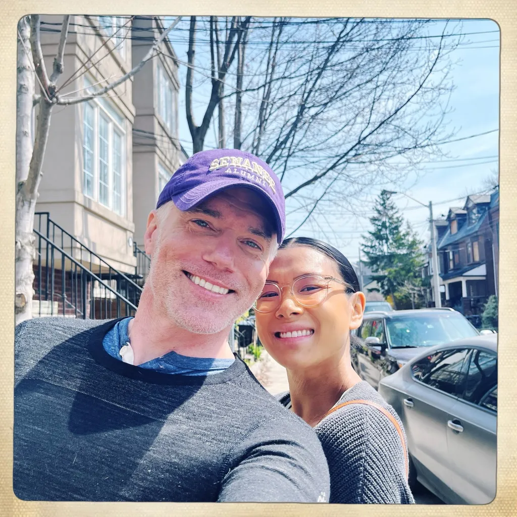 A man and a woman smiling at the camera outdoors, with a street and buildings in the background.