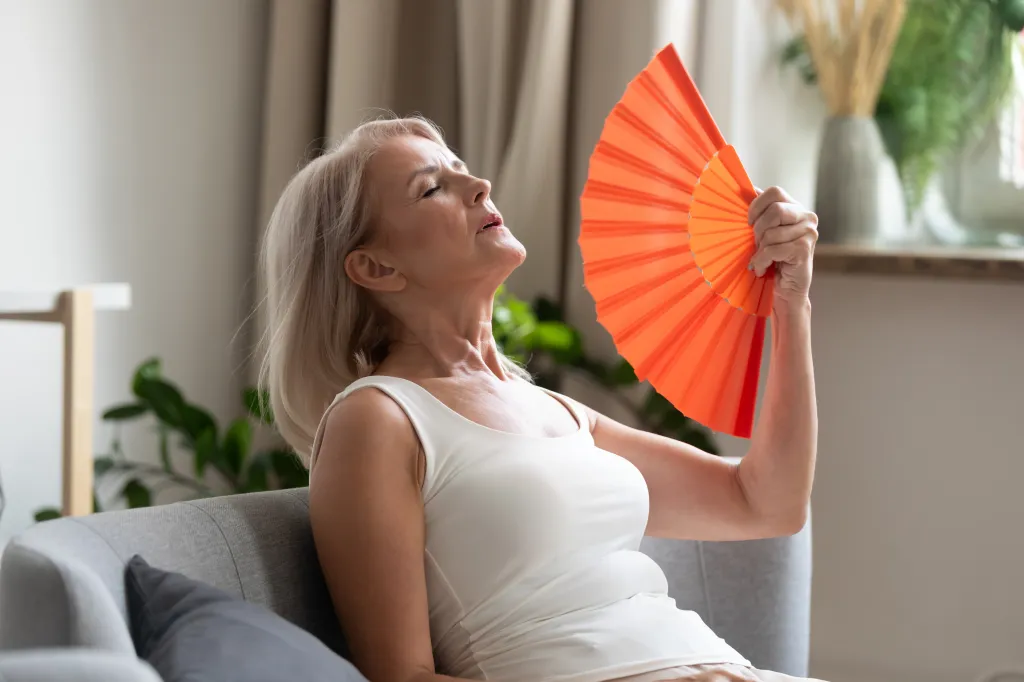 An older woman with blonde hair, wearing a white tank top, sitting on a gray sofa, and using an orange fan to cool herself.