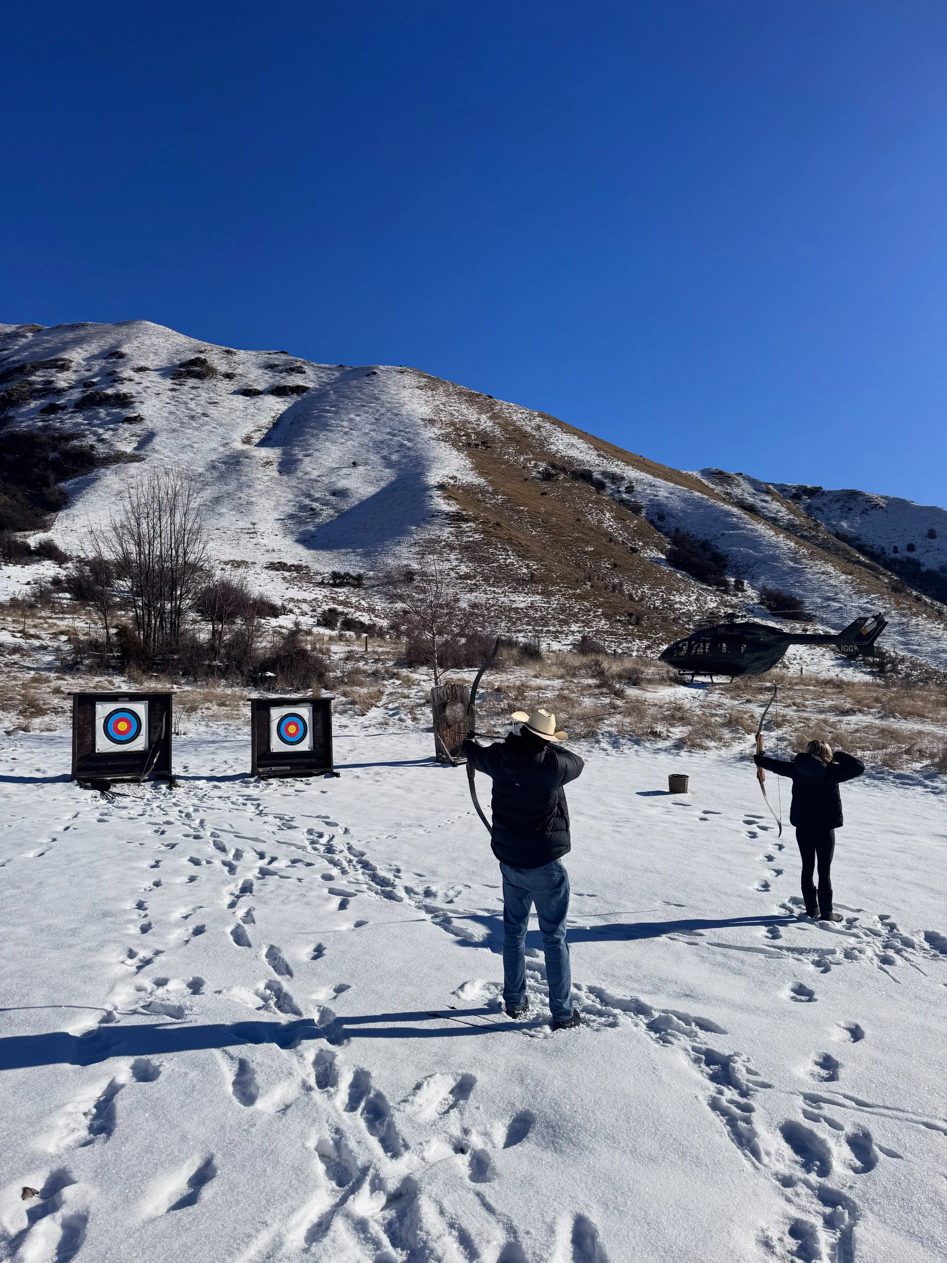 Two people archery target practice in the snow with a helicopter and snowy mountains in the background.
