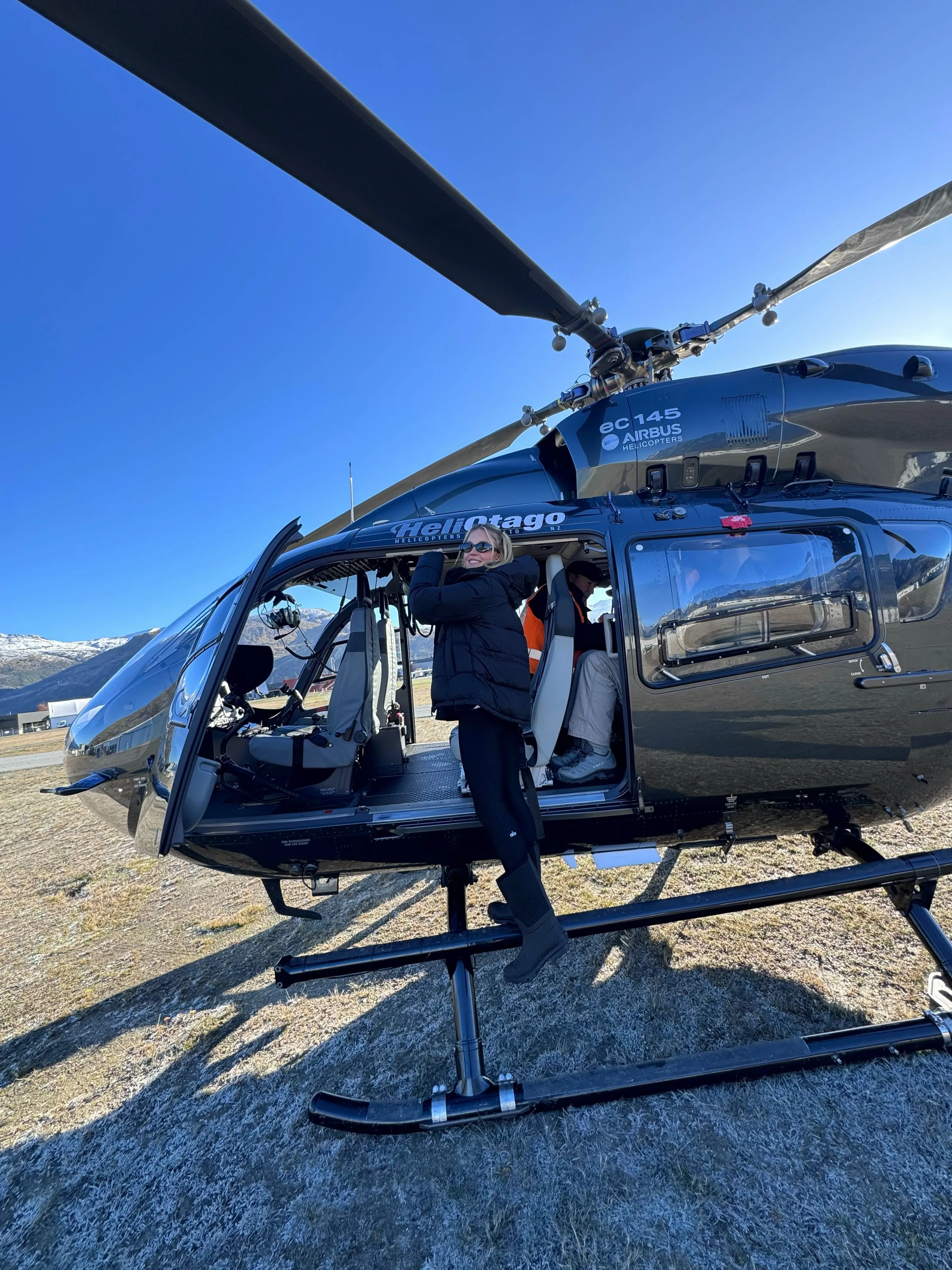Sydney Sweeney posing in front of a dark gray helicopter with a snow-capped mountain in the background.