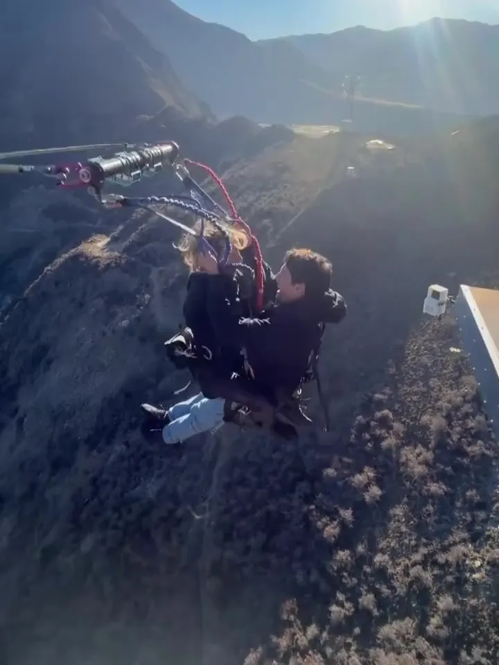 Sydney Sweeney and another person flying through the air, harnessed to a giant swing over a mountain valley.