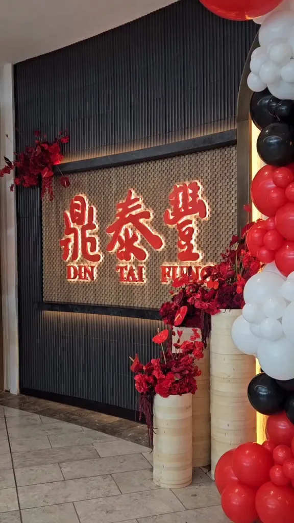 The illuminated Din Tai Fung sign on a textured wall, decorated with red floral arrangements and red, black, and white balloons.
