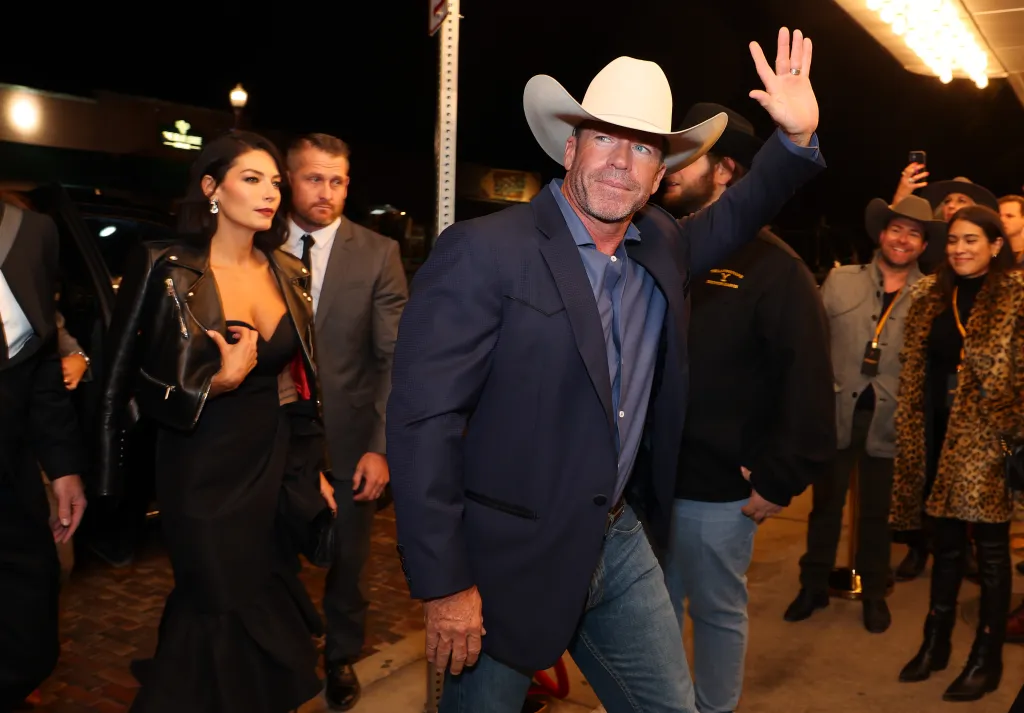 Taylor Sheridan in a cowboy hat and blue suit waves while his wife Nicole Sheridan in a black dress and leather jacket walks beside him at the 