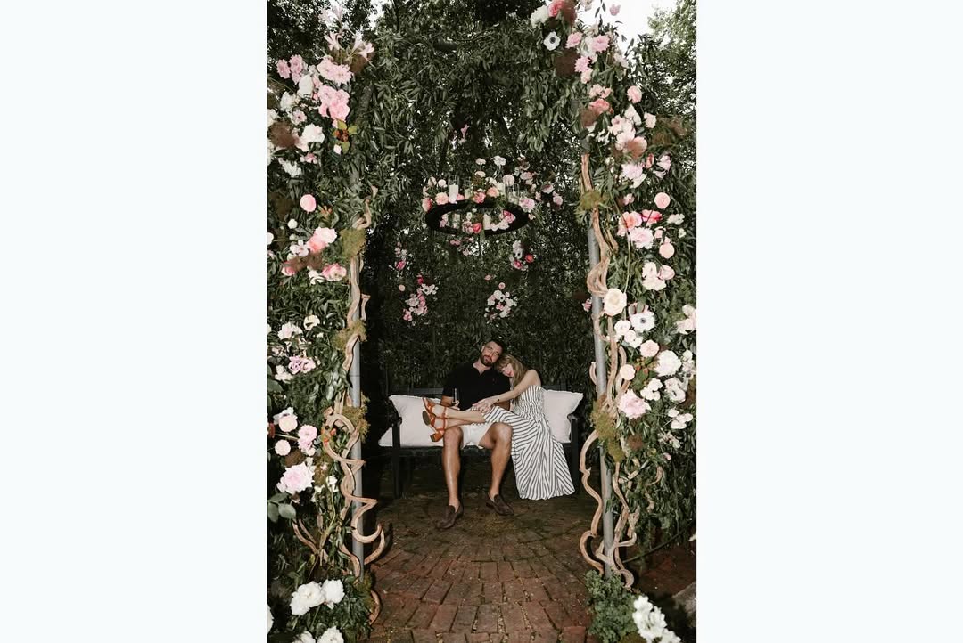 Taylor Swift and Travis Kelce sitting on a swing under a floral archway.