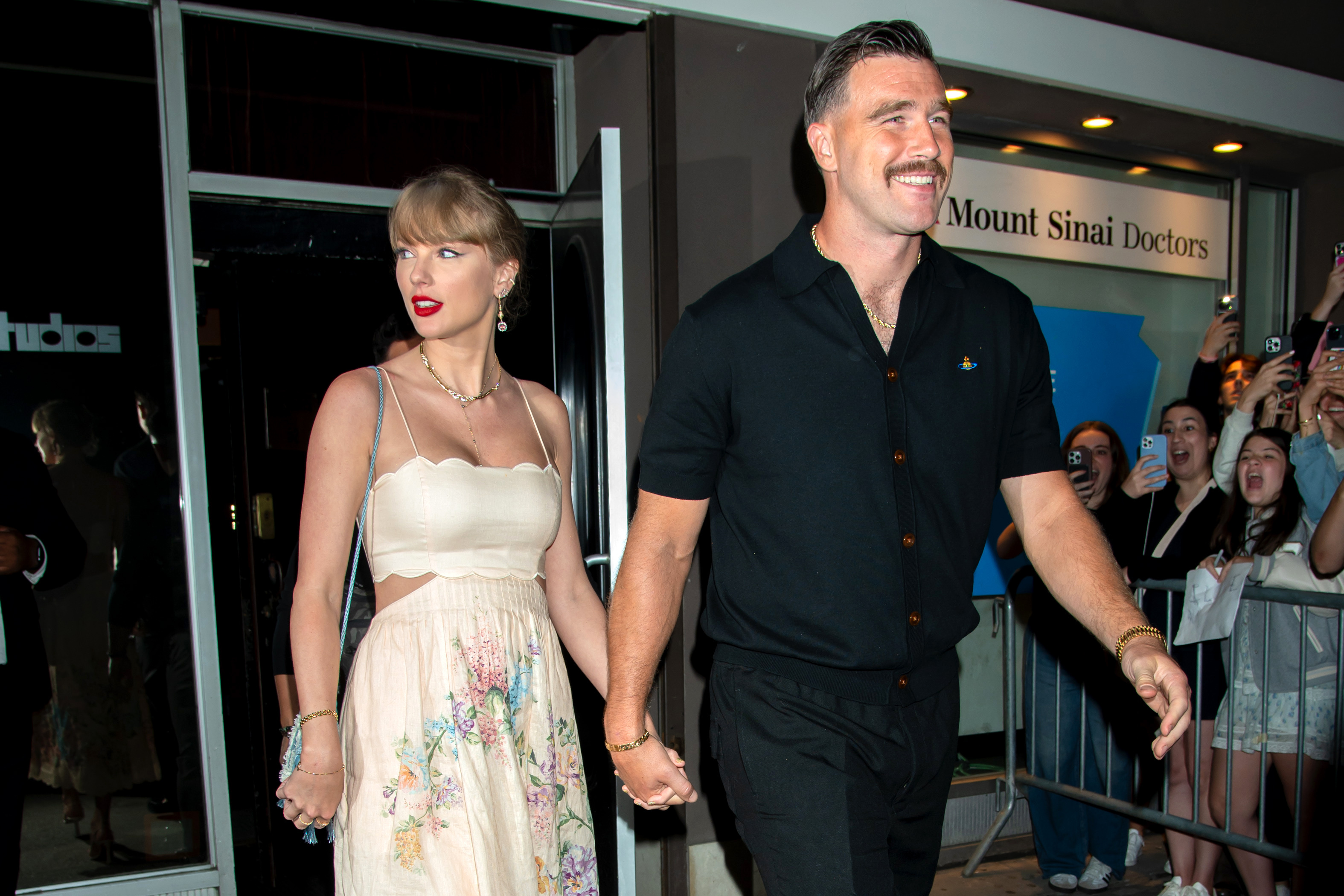Taylor Swift and Travis Kelce holding hands while leaving an event, with fans taking photos in the background.