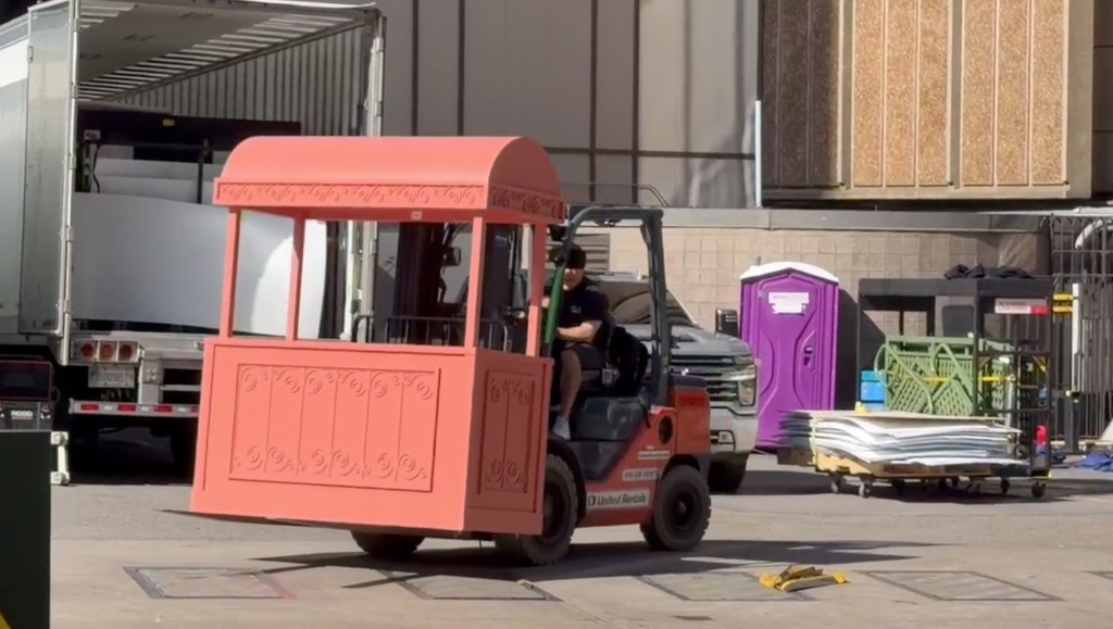 A forklift carrying a red, ornate decoration, possibly a gazebo or a stage, drives near a semi-truck and a purple porta-potty.