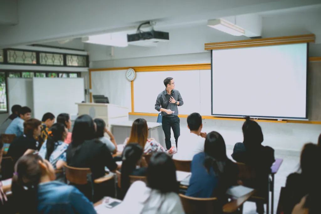 A male teacher giving a lecture to students in a university classroom with a projector screen.
